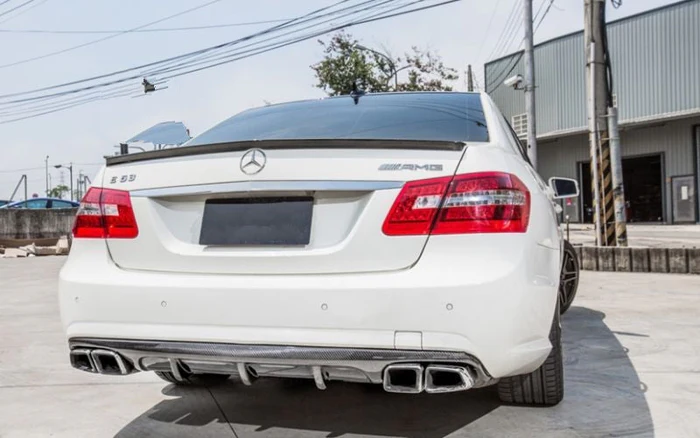 A white Mercedes-Benz E 63 AMG is parked on a concrete surface. It features quad exhausts and is located near a gray industrial building with overhead power lines.