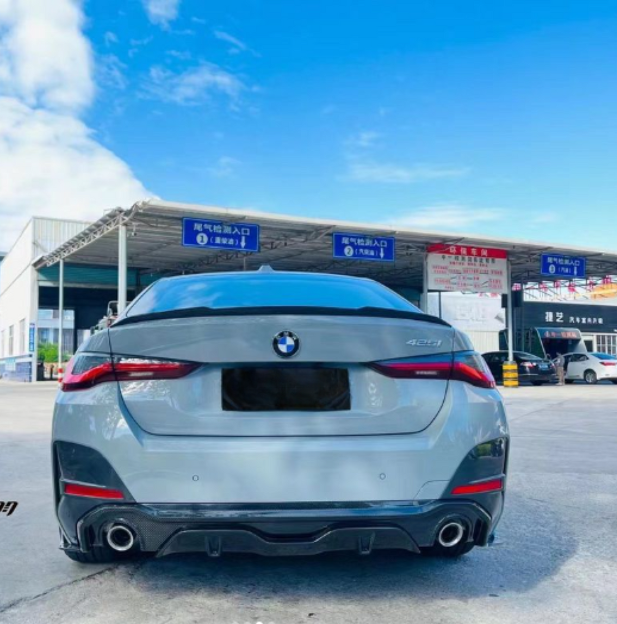 A silver BMW 4-series car is parked in front of an automotive service station. Blue signs in Chinese above indicate the entrance as "尾气检测入口" (Exhaust Inspection Entrance).