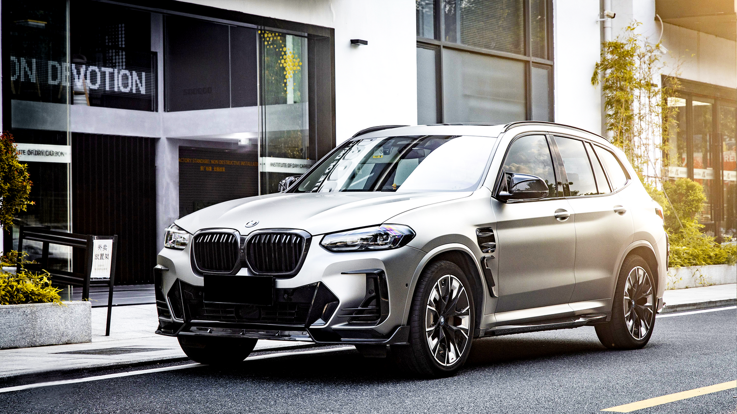 A silver SUV is parked on a city street near a modern building with large windows. Surrounding plants add greenery. Visible text: "INSTITUTE OF DRY CARBON."
