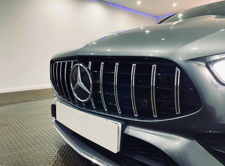 Car front with Mercedes-Benz logo on grille, parked in a lit indoor space with a metallic floor and white walls. A white license plate is attached below the grille.