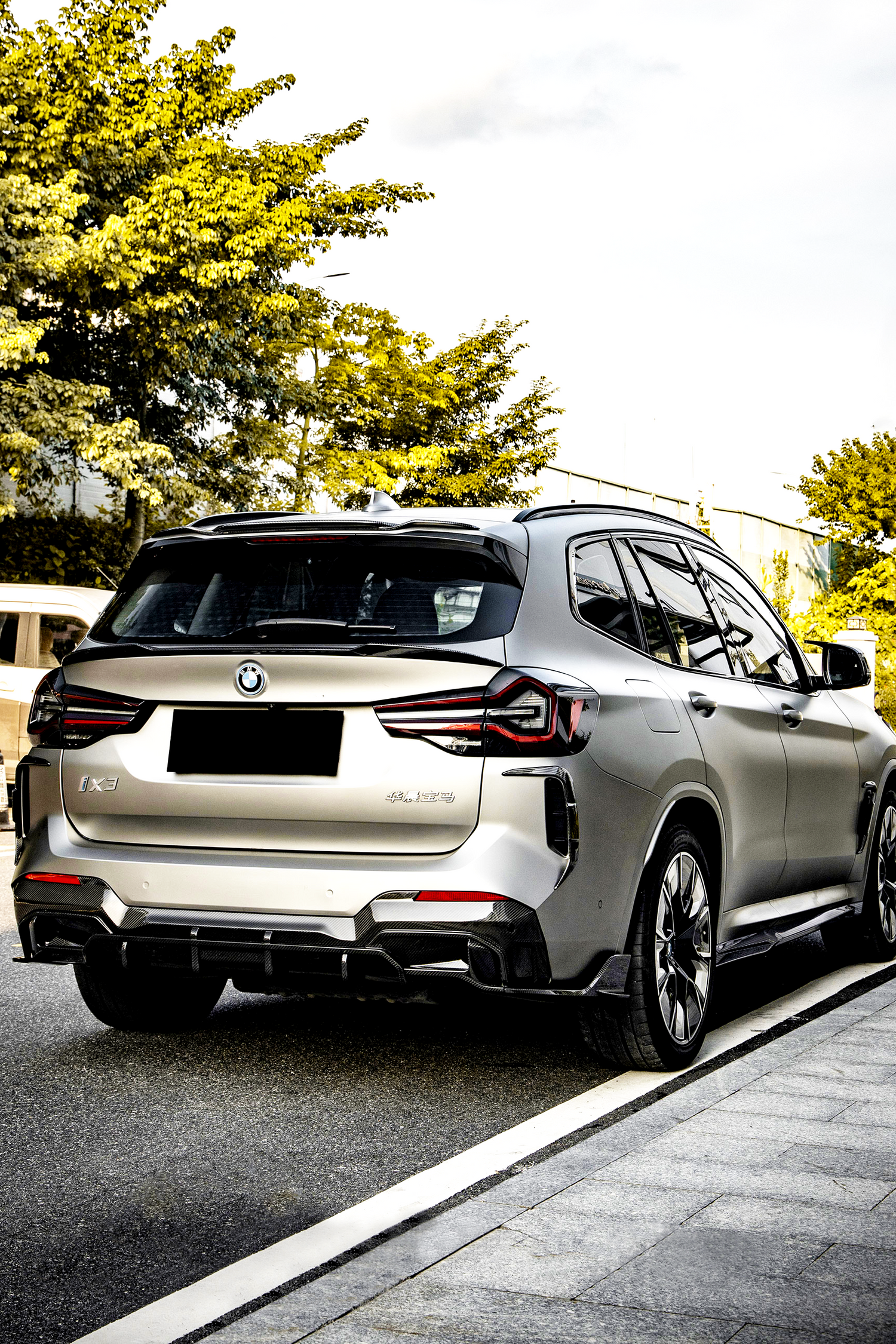 A silver SUV is parked on a road beside a sidewalk, surrounded by green trees. The vehicle has a BMW logo and "iX3" and "Electric" badges visible.