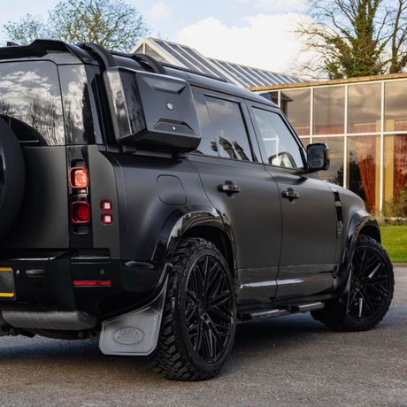 A black SUV parked on a paved surface. The vehicle has large black wheels and a spare tire mounted on the rear. Surrounding environment includes a building with glass windows and trees in the background.