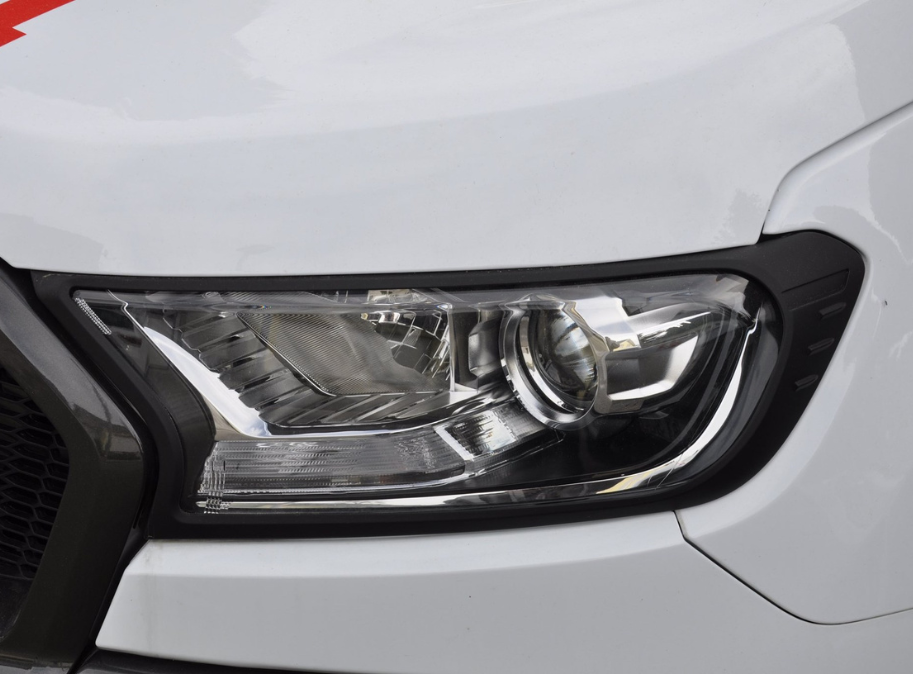 A close-up view of a car headlight, featuring a sleek, angular design with reflective surfaces, set within a white vehicle body. The headlight is bordered by black trim.
