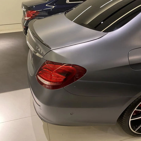 A gray luxury car's rear with "E 43" badge; parked beside another blue car inside a showroom with reflective flooring.