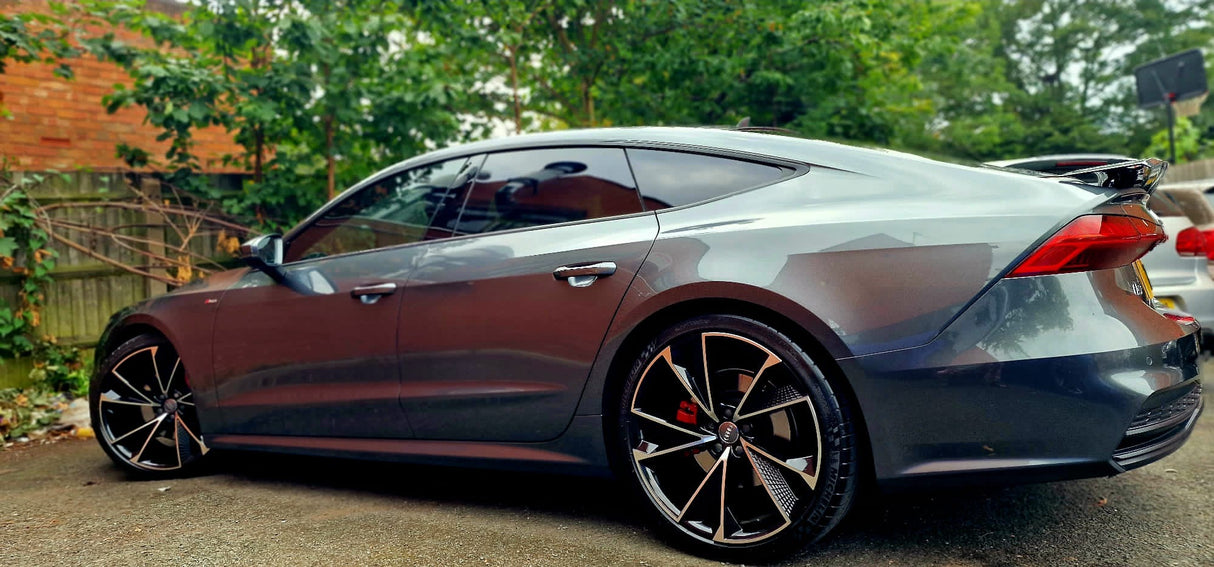 A sleek, dark gray car is parked, showcasing its modern, intricate rims. It's set in a driveway bordered by a wooden fence and green foliage.