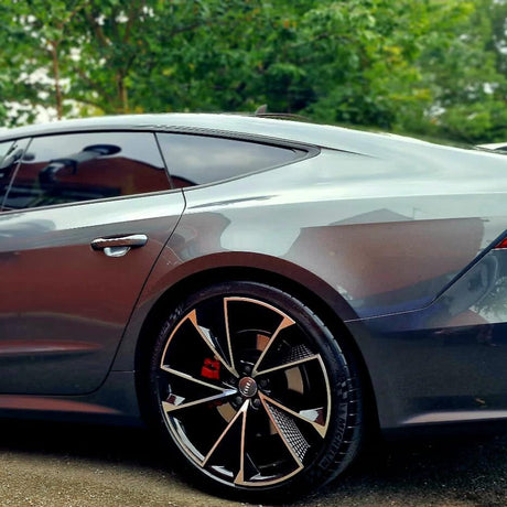 A sleek gray sports car is parked, showcasing its polished body and distinctive alloy wheels. Surrounding trees and greenery slightly blur in the background, emphasizing the car's design.