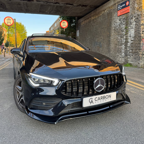 Car: A black Mercedes-Benz, stationary, with a "CARBON ACCENTS" license plate. Context: Parked under a bridge with brick walls, surrounded by "No Cycling" signs and a nearby street with trees and buildings.