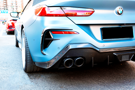 A blue BMW car is parked on a city street. It features dual exhausts and carbon fiber detailing, with other vehicles visible in the background.