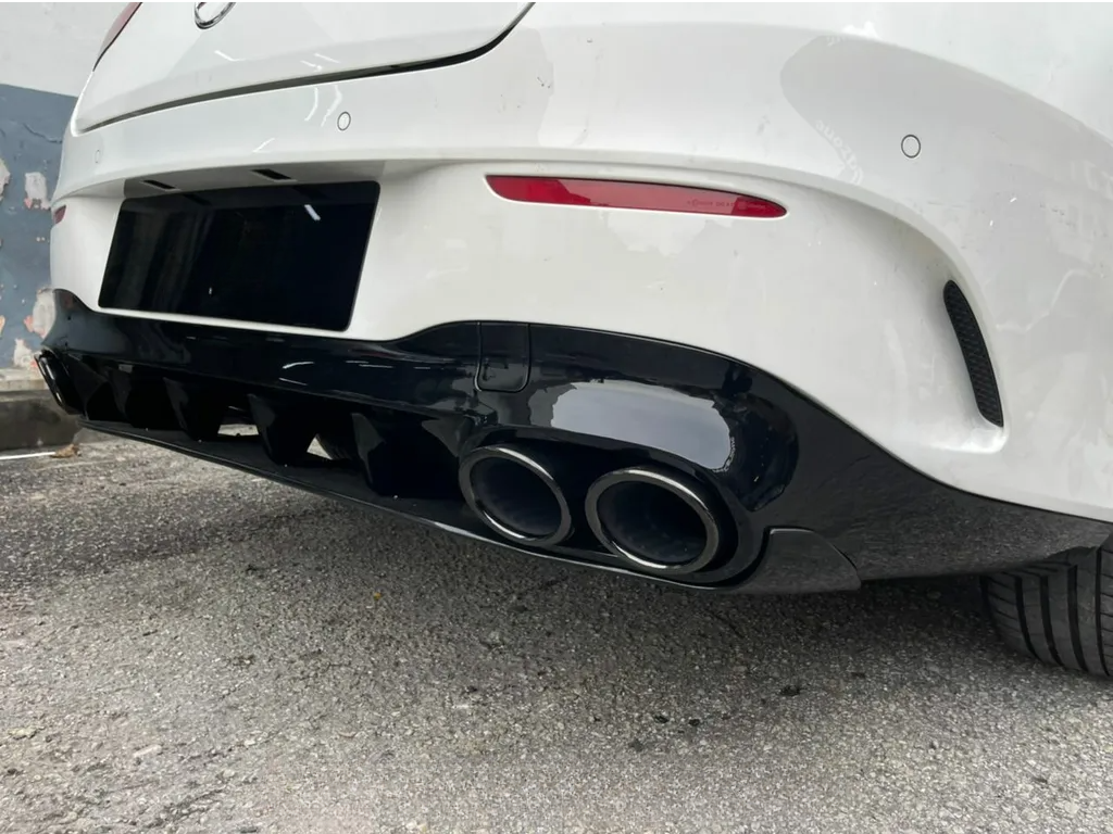 A car's rear bumper displays dual exhaust pipes, parked on a rough asphalt surface. The white vehicle features a black diffuser and a red reflector amid an urban environment.