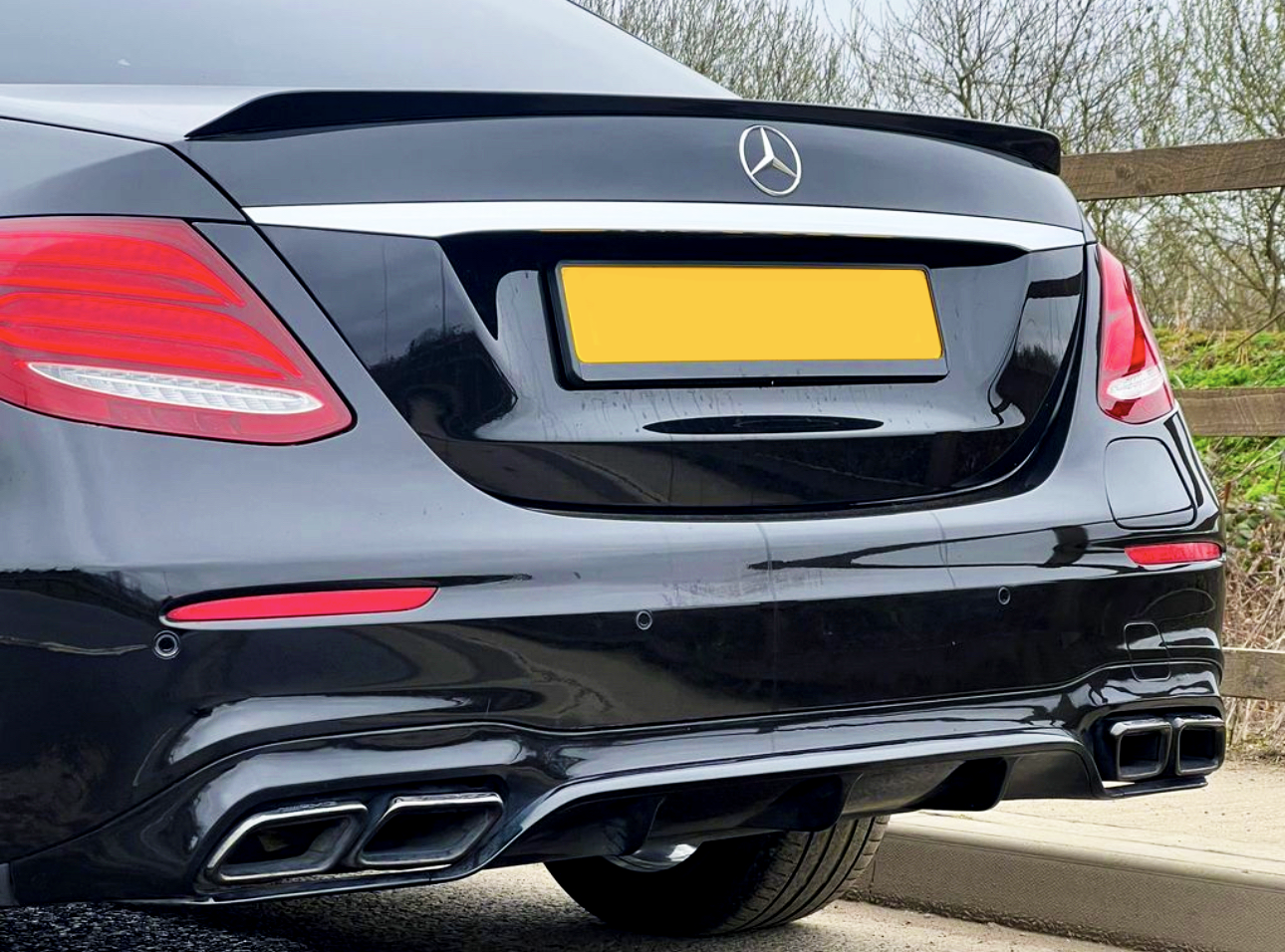 A black luxury car is parked by a wooden fence, showcasing its rear with red taillights and dual exhausts. The car has a yellow license plate and a silver emblem.