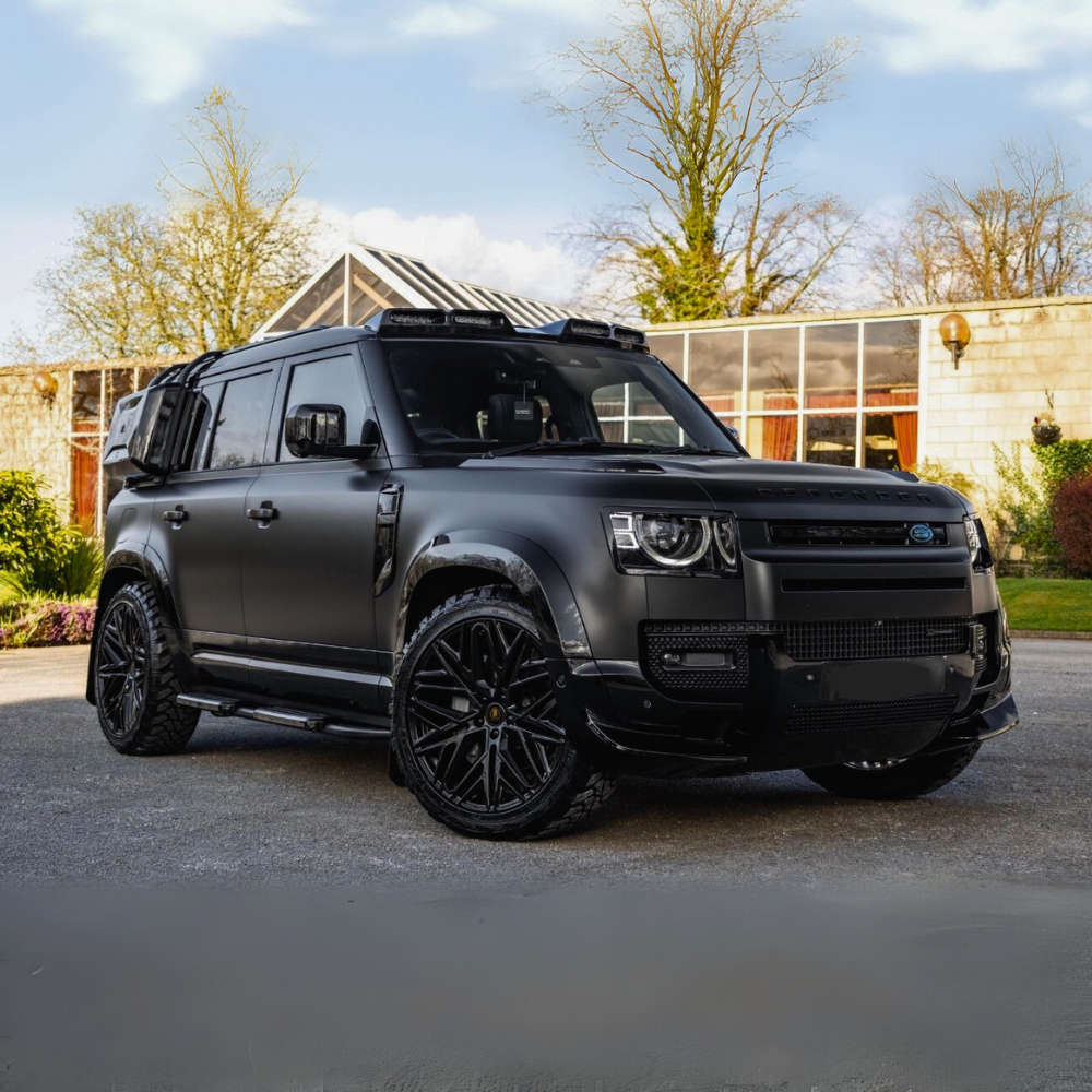 A matte black SUV is parked on a driveway in front of a house with large windows. Surrounding the vehicle are trees and bushes, under a clear blue sky.