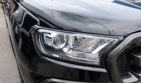 A sleek black vehicle's headlight assembly, reflecting light, is prominently displayed. It is parked on a concrete surface, with a cloudy sky reflecting on the car's surface.