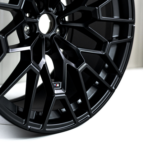 Black car alloy wheel with intricate spokes is displayed against a neutral background. A small emblem with an "M" on it is visible in the center section.