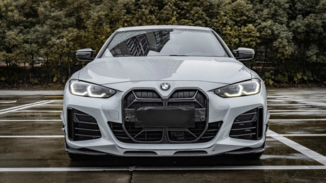 A white car is parked on a wet, empty parking lot. Its headlights are on, and trees form the background. The BMW logo is visible on the hood.