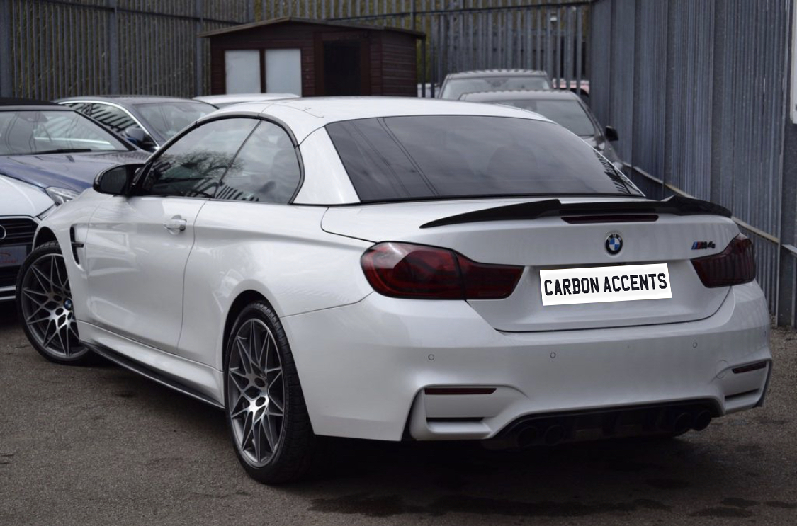 A white BMW M4 is parked on a lot with other cars, bordered by a fence. The license plate reads "CARBON ACCENTS." A small wooden shed is in the background.