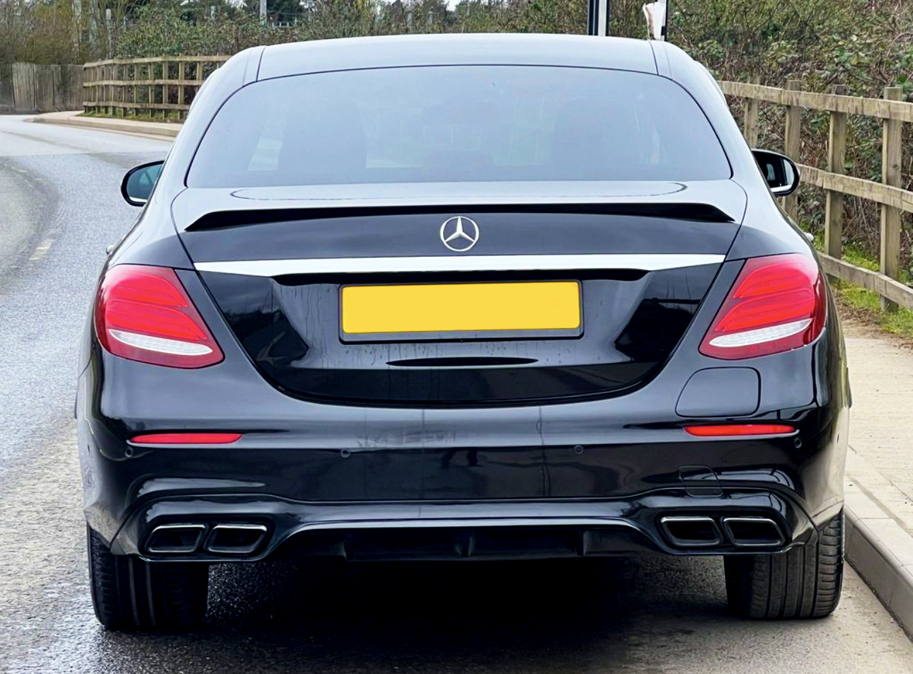 A black car is parked on a quiet road, aligned next to a wooden fence. The license plate is yellow, and a Mercedes-Benz logo is visible on the trunk.