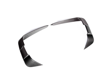 Two carbon fiber trim pieces, angled and sleek, lie on a plain white background, resembling modern automotive design elements, possibly for enhancing vehicle aerodynamics or styling.