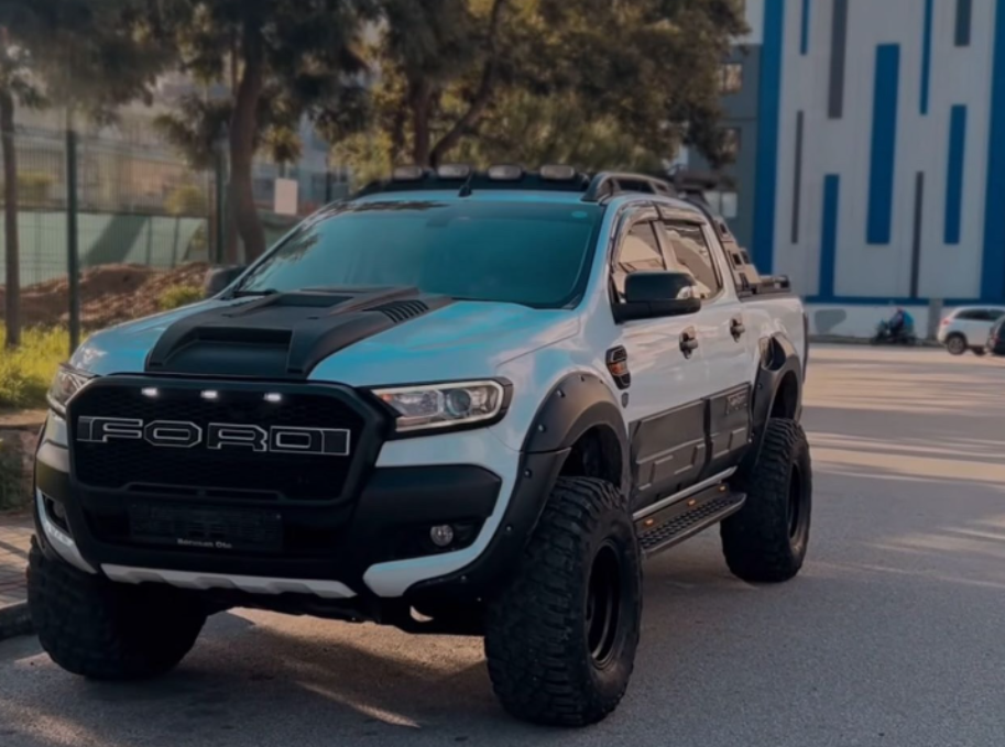Pickup truck parked on a street, featuring large off-road tires and a custom grille with "FORD" lettering. Surrounding trees and modern buildings provide an urban backdrop.
