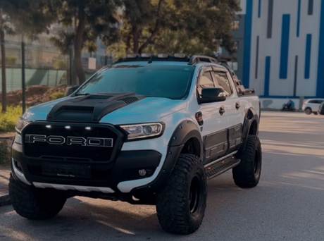 Pickup truck parked on a street, featuring large off-road tires and a custom grille with "FORD" lettering. Surrounding trees and modern buildings provide an urban backdrop.