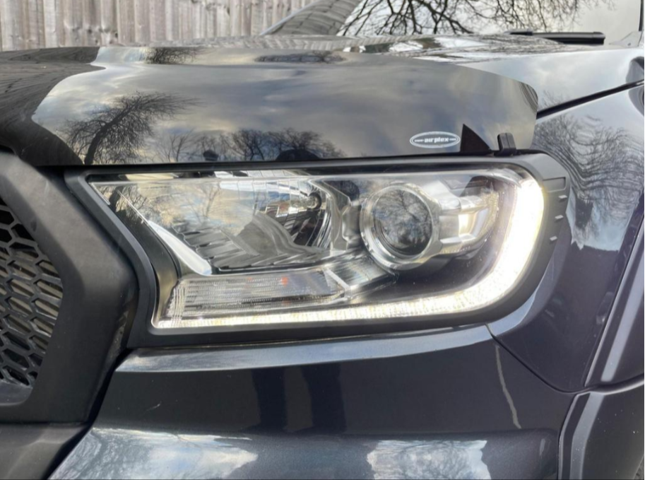 A car's headlight, reflecting nearby trees, is illuminated in daylight. It features a sleek design, situated next to a dark grille, with an "arplex" logo visible on the lens.