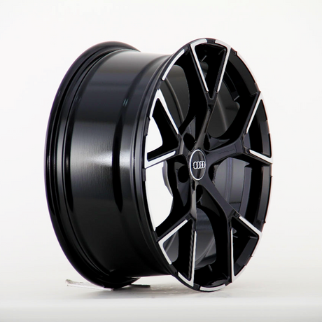 A black alloy wheel with a multi-spoke design lies on its side, showcasing the Audi logo at the center. The background is a plain white surface.