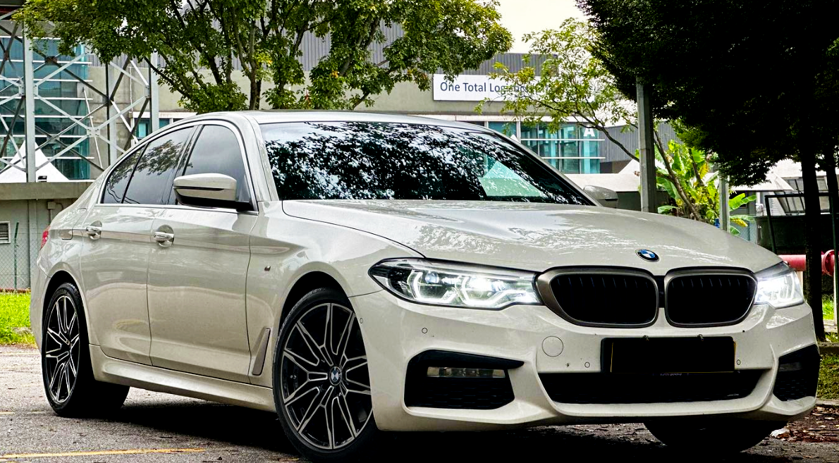 A white BMW sedan is parked on a road, surrounded by trees and buildings. A sign in the background reads "One Total Logistics."