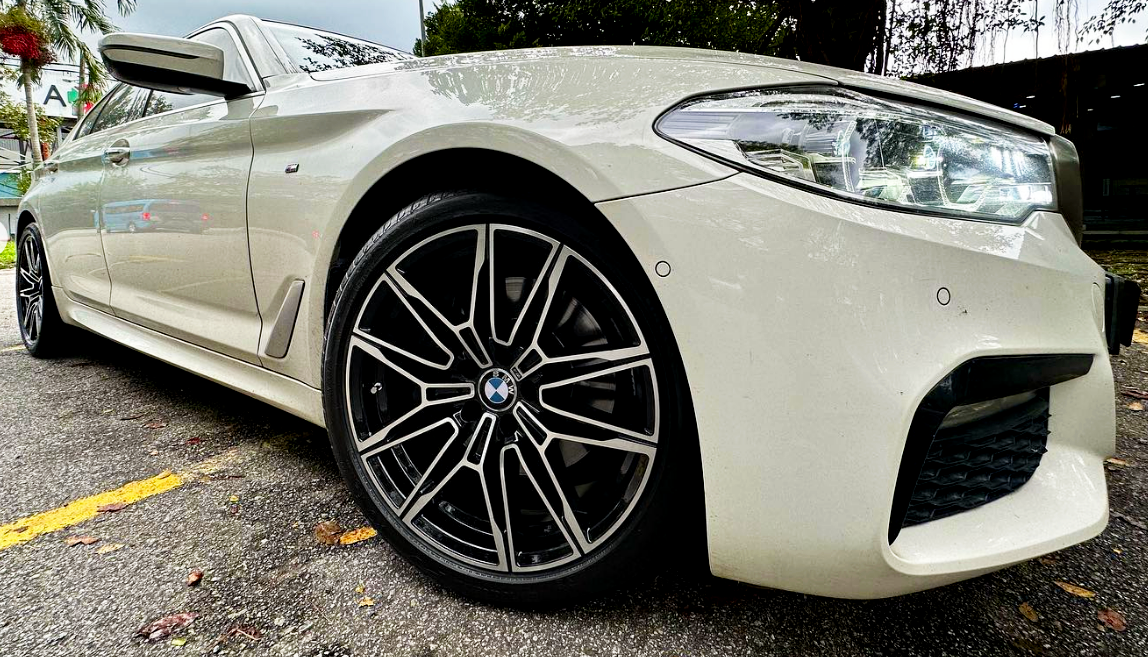 A white BMW car is parked on a street, showcasing its sleek front design and distinctive alloy wheels. Surrounding trees and signs create a busy urban environment.
