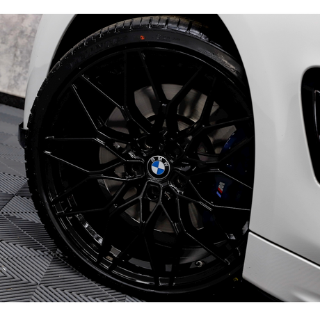 A black, multi-spoke BMW wheel is stationary, mounted on a white car. The vehicle rests on a textured black rubber mat, set indoors against a gray wall.