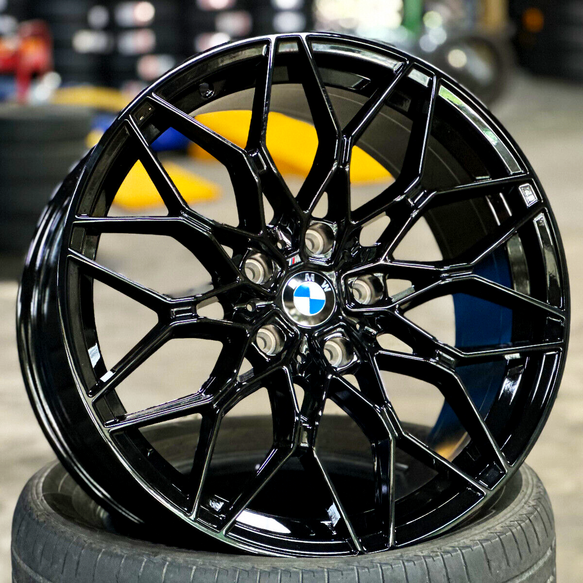 A glossy black BMW wheel rests on a tire, displaying intricate spoke design. It is set in a workshop environment with blurred tools and equipment in the background.