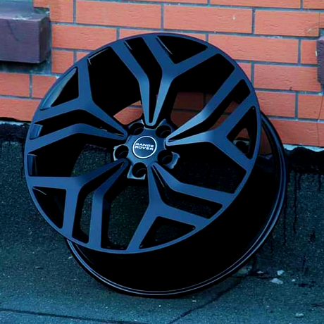 A black alloy wheel rests against a brick wall. The wheel features angular spokes and a central hubcap with the text "Range Rover" in white letters.