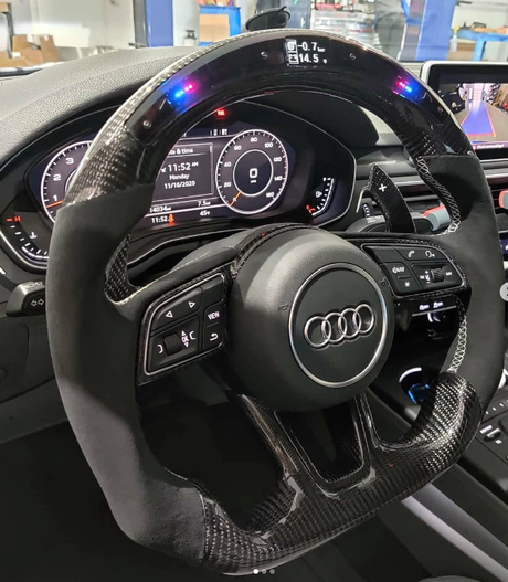 A carbon fiber car steering wheel with buttons is centered in the dashboard of an Audi. Digital displays show "11:52 AM," "11/16/2020," speed, and other metrics.