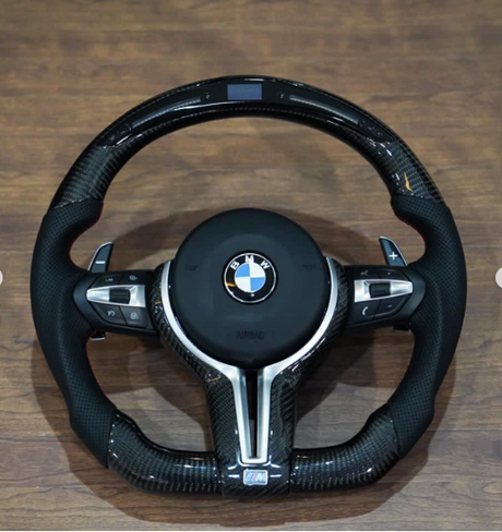 The object is a BMW car steering wheel with paddle shifters and control buttons, placed on a wooden surface. Text visible includes "BMW," "AIRBAG," and "M."