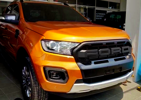 An orange pickup truck is parked indoors, showcasing a prominent front grille with "FORD" branding. The truck is situated in a well-lit showroom with signage in the background.