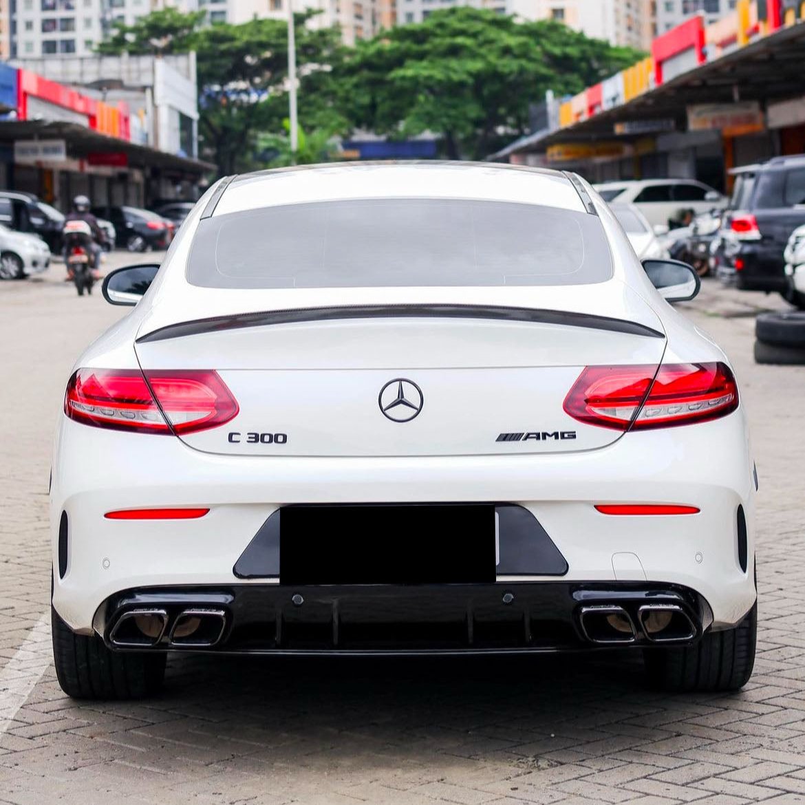 A white Mercedes-Benz C 300 AMG is parked on a cobblestone area, surrounded by parked vehicles and buildings, with greenery in the background.