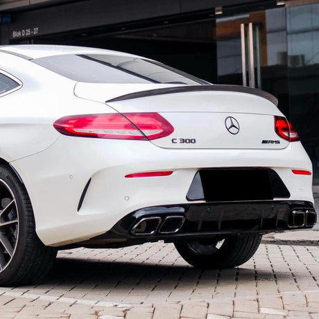 A white Mercedes-Benz C 300 AMG parked on a cobblestone surface, displaying dual exhausts under its rear bumper, near modern building. Text: “C 300” and “AMG” badges.