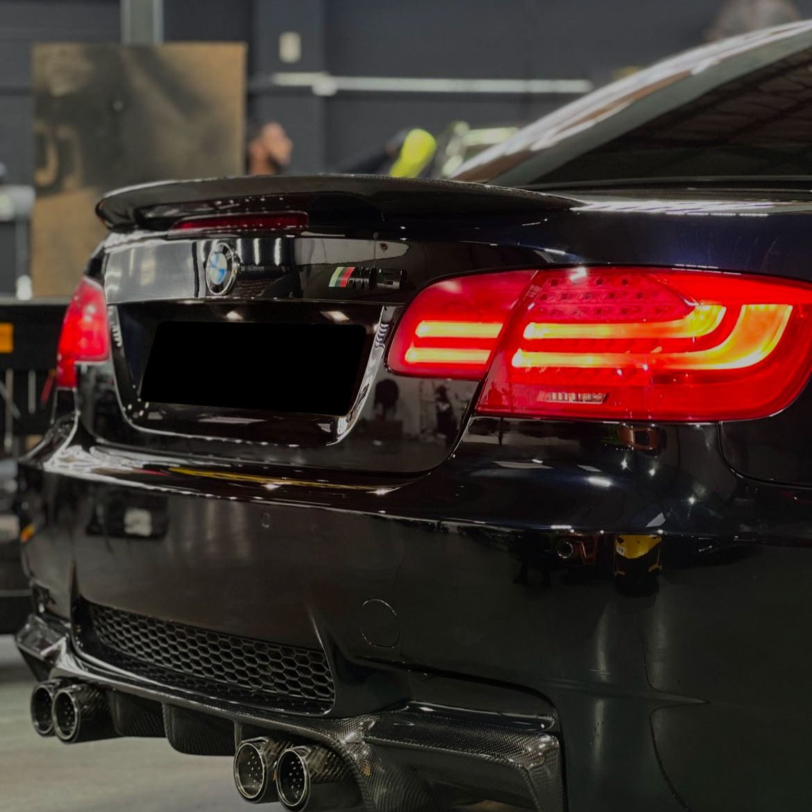 A black BMW M3 with illuminated red taillights is parked indoors. The dual exhaust and carbon fiber bumper highlight its sporty design. A blurred workshop setting surrounds the car.