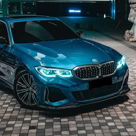 A blue car with illuminated headlights is parked on a checkered pavement beside a building and a statue, under ambient lighting.