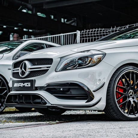 A white Mercedes-Benz AMG car is parked on a concrete surface, featuring a prominent grille and sleek wheel design. A fence and an industrial structure are in the background.