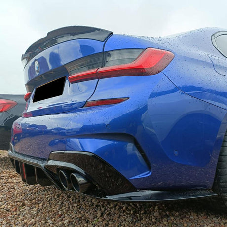 A blue sports car with visible exhaust pipes and a rear spoiler is parked on gravel, showing rain droplets on its surface under an overcast sky.