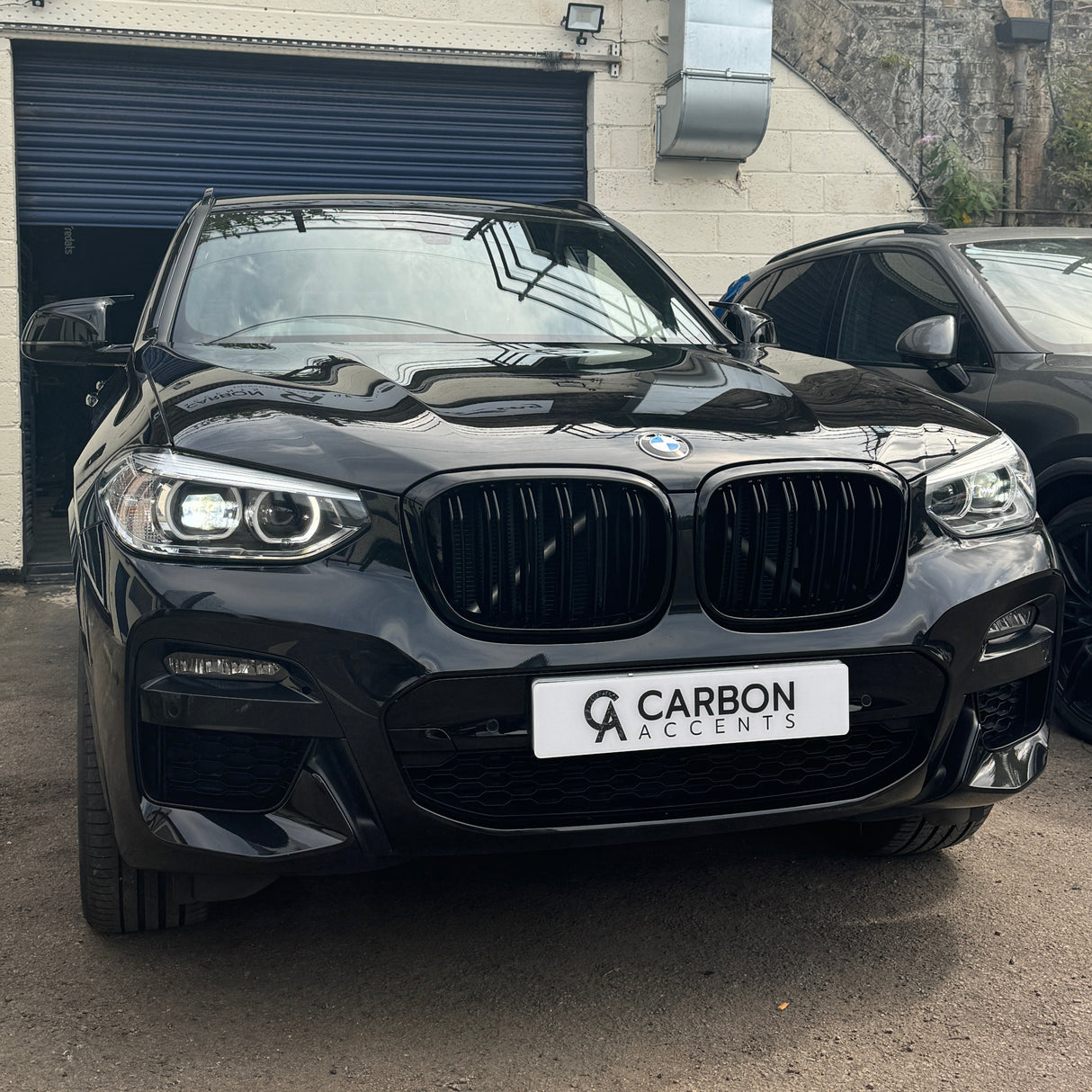 A black BMW SUV is parked in front of a garage with a blue door. The license plate reads "CARBON ACCENTS." Another vehicle is partially visible beside it.