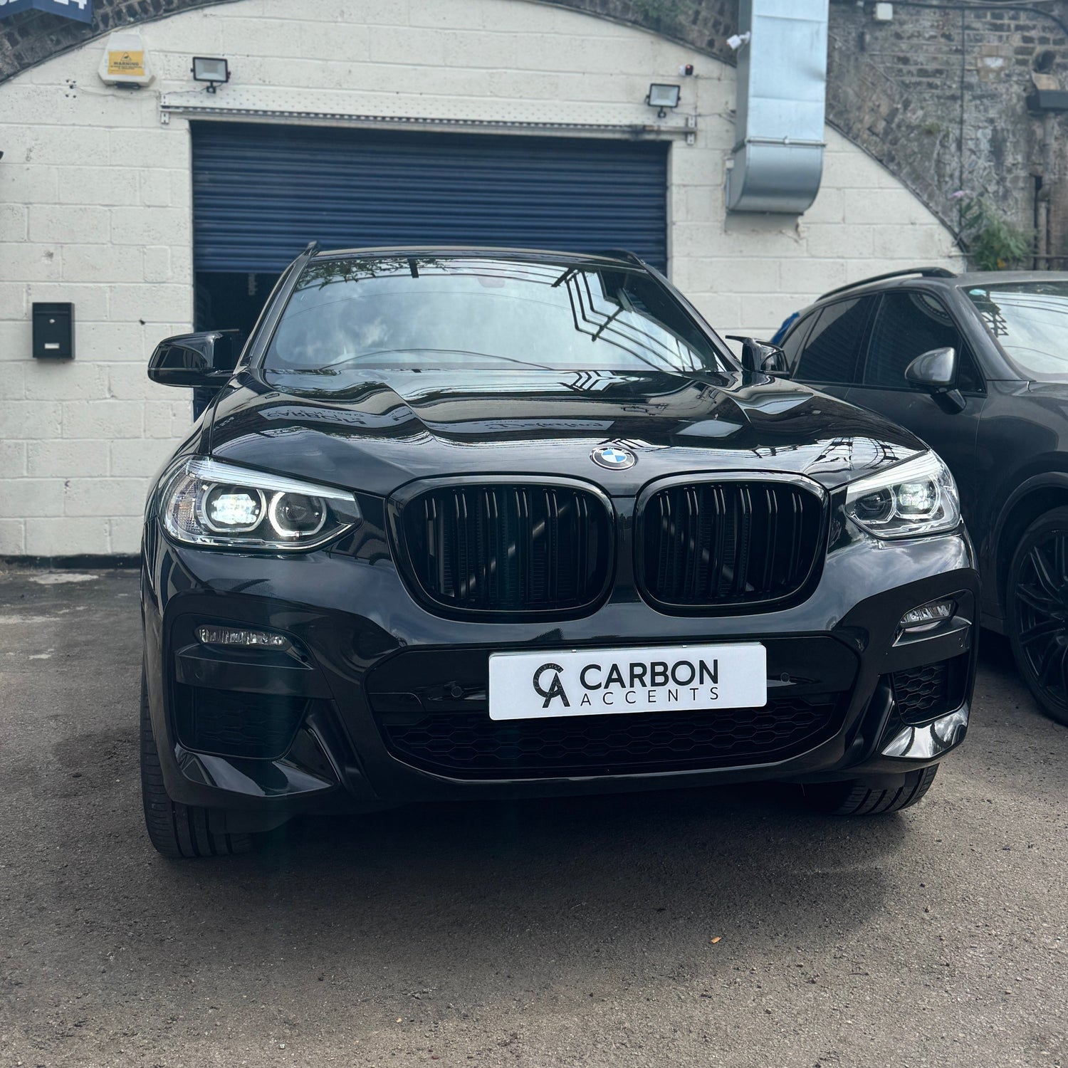 A black BMW parked in front of a garage, displaying a "CARBON ACCENTS" license plate. The setting includes a white brick wall with a closed blue door.