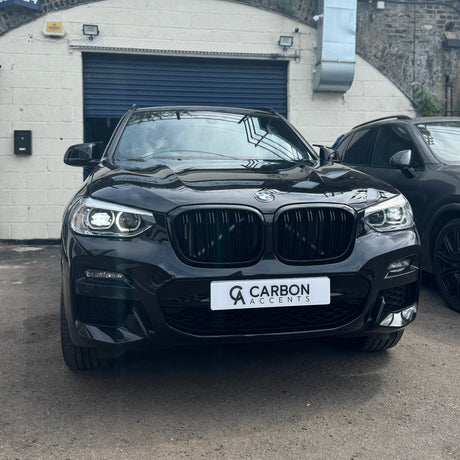 A black BMW parked in front of a garage, displaying a "CARBON ACCENTS" license plate. The setting includes a white brick wall with a closed blue door.
