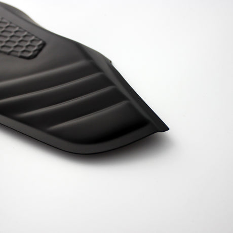 A black, ribbed car floor mat corner with a textured rectangular section rests on a smooth white surface, showing its contoured edges.