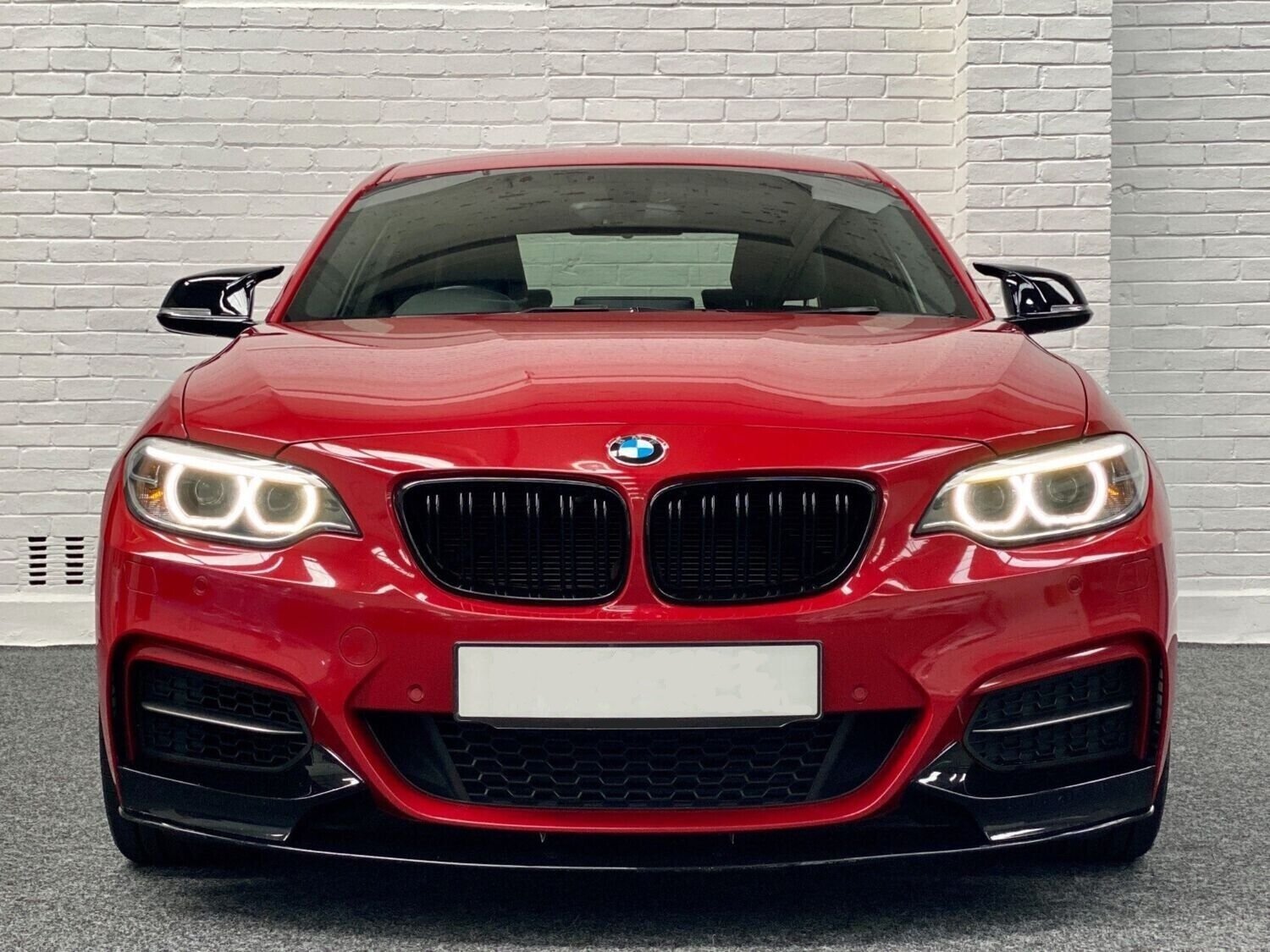 A red BMW car, headlights on, is parked indoors against a white brick wall backdrop.