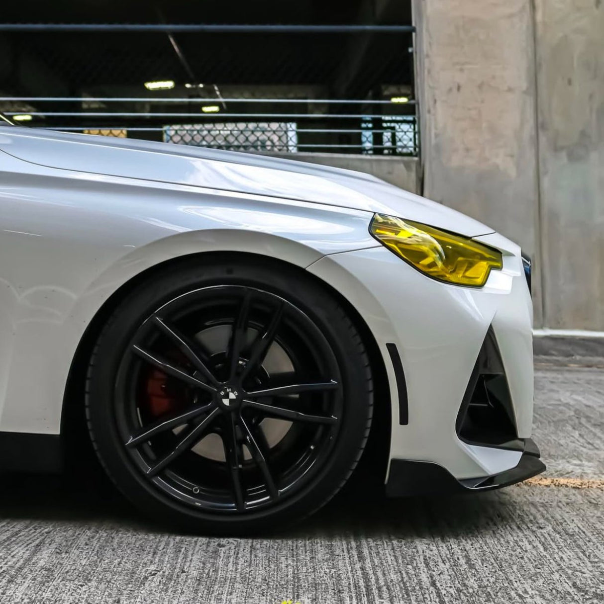 A white car with black wheels is parked on concrete. The front headlight is yellow. The setting is an urban parking area with a metal fence and concrete walls.