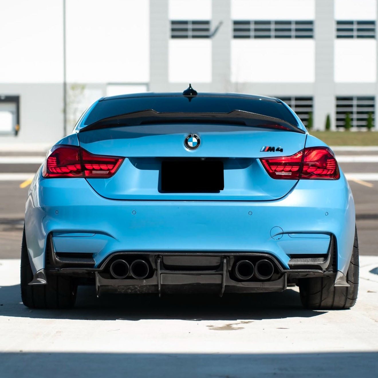 A blue BMW M4 is parked with noticeable quad exhausts and red taillights, situated in a sunny urban parking lot with a modern building backdrop.