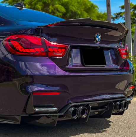 A BMW sports car with a shiny purple finish, features prominent dual exhausts and red taillights, parked on a sunny day with lush greenery and palm trees as the backdrop.