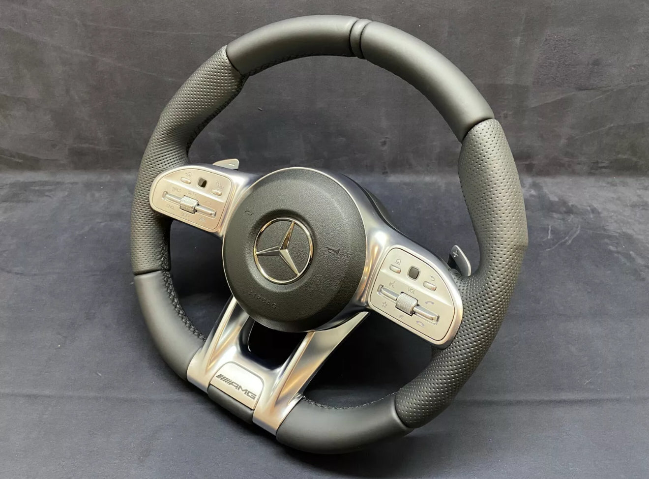 The steering wheel features a central Mercedes logo and silver controls on either side, set against a black, textured grip, and displayed on a plain, dark surface.