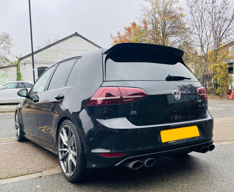 A black hatchback car is parked on a street near autumn trees and a warehouse. It features dual exhausts and alloy wheels, with a visible "VW" logo on the rear.
