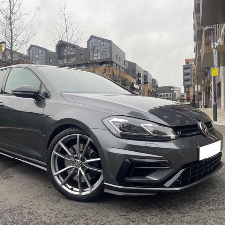 A gray car is parked on a street with modern buildings and barren trees in the background, under an overcast sky.